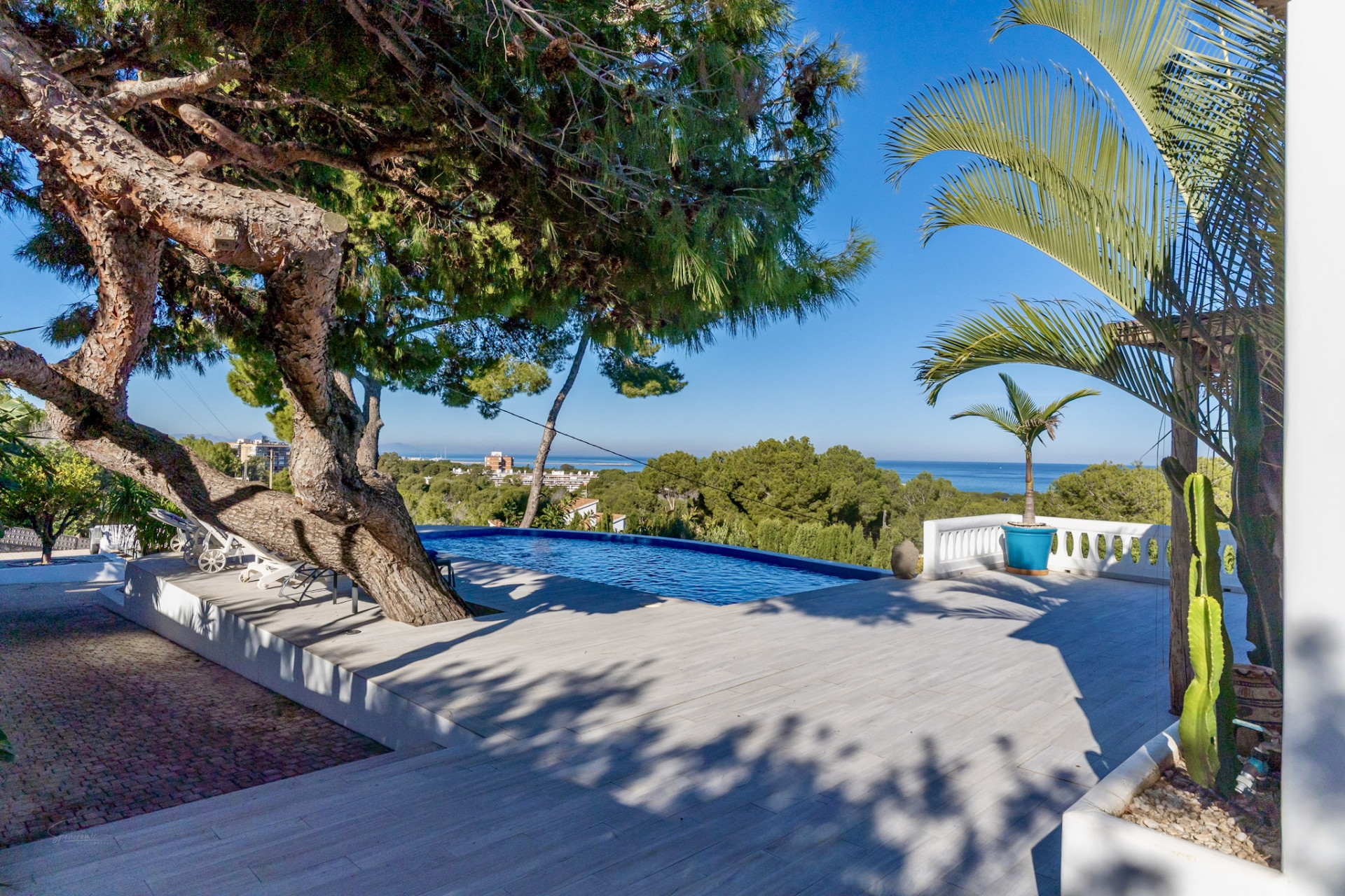 Terraza con piscina infinita de azulejos claros, enmarcada por un pino maduro y balaustrada blanca. Ofrece vistas panorámicas al mar y tumbonas. Terraza con piscina infinita de azulejos claros, enmarcada por un pino maduro y balaustrada blanca. Ofrece vistas panorámicas al mar y tumbonas.