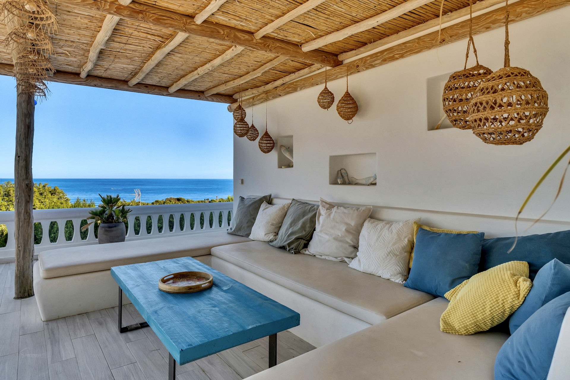 Terraza cubierta con asientos integrados, cojines variados y una mesa de madera azul. El techo de caña natural complementa las vistas panorámicas al mar Mediterráneo. Terraza cubierta con asientos integrados, cojines variados y una mesa de madera azul. El techo de caña natural complementa las vistas panorámicas al mar Mediterráneo.