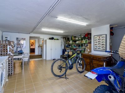 Amplia sala de servicio con suelo de baldosas claras, equipada con lavadora, estanterías, y espacio para una bicicleta eléctrica y una moto, bajo iluminación fluorescente.