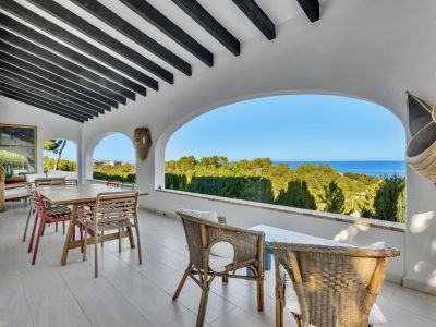 Amplia terraza cubierta con vigas de madera oscura y arcos, ofreciendo vistas panorámicas a la vegetación y al mar Mediterráneo. Incluye comedor y zona de asientos de mimbre.