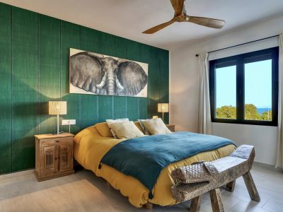 Amplio dormitorio con pared de acento verde texturizada, cuadro de elefante y banco de madera tallada. Ventana con vistas lejanas al mar y ventilador de techo de madera.