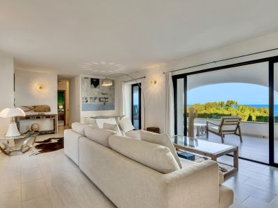 Amplio salón con sofá beige, suelo de baldosas claras y acceso directo a una terraza con vistas panorámicas al mar y vegetación exuberante. Incluye mesa de centro moderna y arte mural.
