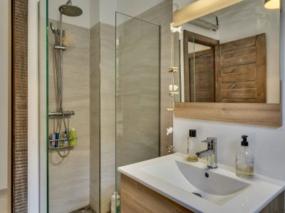 Baño con ducha moderna de mampara de cristal, azulejos claros y suelo de guijarros. Lavabo con mueble de madera clara y espejo con marco a juego.