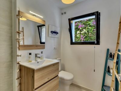 Baño con tocador de madera clara, espejo grande con iluminación LED integrada y ventana con vista a vibrantes buganvillas rosas. Incluye inodoro y un móvil decorativo de peces de madera.