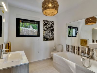 Baño luminoso con bañera, lavabo de diseño y ventanas que ofrecen vistas a la vegetación exterior. Destacan la lámpara de mimbre y el cuadro de cebras, complementados por azulejos claros y amplios espejos.