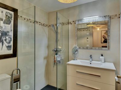 Baño moderno con ducha de cristal, azulejos beige y cenefa decorativa. Incluye un mueble de lavabo blanco con espejo de marco rústico y luz integrada.
