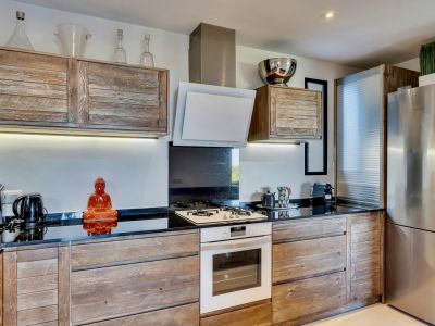 Cocina contemporánea con gabinetes de madera rústica, encimeras negras y horno blanco integrado con placa de gas. La iluminación bajo los armarios realza el salpicadero oscuro y el frigorífico de acero inoxidable.