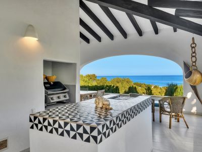Cocina exterior bajo pérgola con encimera de azulejos geométricos en blanco y negro, barbacoa integrada y vistas panorámicas al mar Mediterráneo a través de un arco.