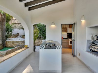 Cocina exterior cubierta con isla central de azulejos decorativos y barbacoa integrada, complementada por un área de parrilla en nicho y vistas al jardín con palmeras.