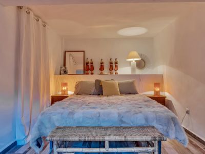 Dormitorio con cama de diseño, cabecero con estante decorado con figuras y lámparas. Banco de ratán y cortinas blancas complementan el ambiente cálido.