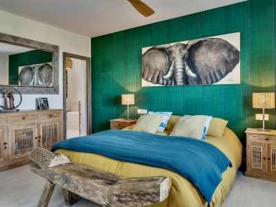 Dormitorio con una llamativa pared de acento verde texturizado, realzada por un cuadro de elefante y mobiliario de madera natural, incluyendo un banco tallado y espejo con marco. Ventilador de techo.