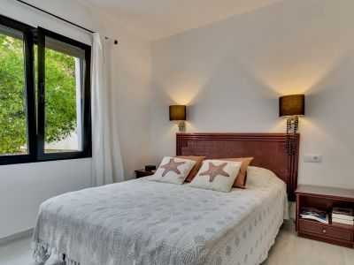 Dormitorio luminoso con cama doble, cabecero de madera oscura y almohadas con estrellas de mar. La ventana abierta con marcos negros ofrece vistas a la vegetación exterior.