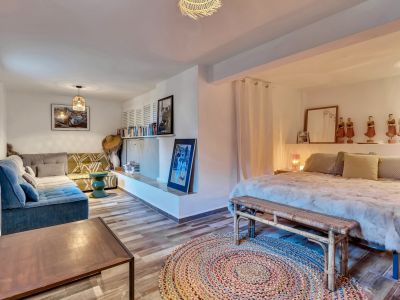 Dormitorio principal con área de estar integrada, suelo de baldosas imitación madera y alfombra redonda. Incluye cama con banco de mimbre, sofá esquinero y estantería con arte.