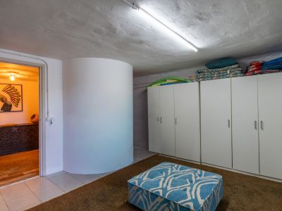 Espacio práctico con armarios blancos para almacenamiento, techo texturizado con luz fluorescente y alfombra marrón. Un puf azul estampado y una puerta abierta revelan la cálida decoración de una sala adyacente.