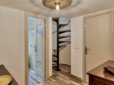 Pasillo con una distintiva escalera de caracol de madera y metal negro, complementado por un suelo de baldosas efecto madera. Se observan dos puertas, una de ellas abierta a un baño, y una consola de madera oscura.