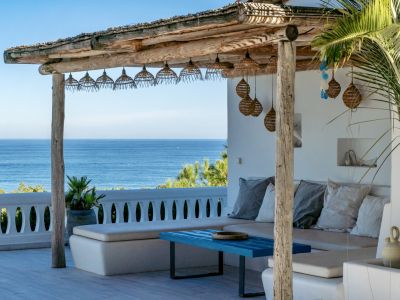 Pérgola de madera rústica con iluminación colgante, ofreciendo un espacio de descanso con asientos blancos integrados y una mesa azul, todo con vistas panorámicas al mar.
