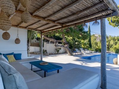 Pérgola rústica con techo de caña y lámparas colgantes de mimbre sobre un sofá exterior con cojines. La zona de piscina y vegetación exuberante se extiende al fondo.