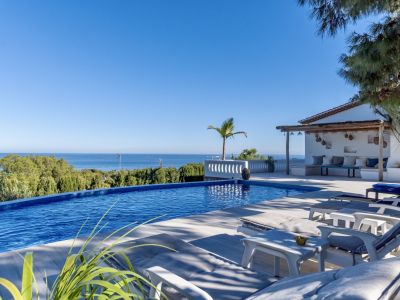 Piscina infinita con vistas panorámicas al mar, rodeada de tumbonas y una zona de asientos rústica bajo pérgola.