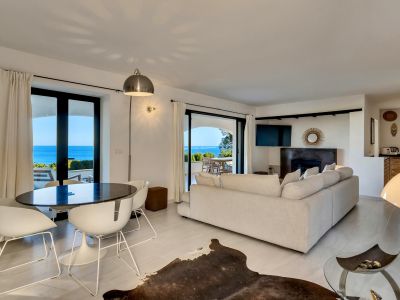 Salón comedor de planta abierta con mesa ovalada oscura, sillas blancas y sofá claro, que se abre a una terraza con vistas panorámicas al mar.