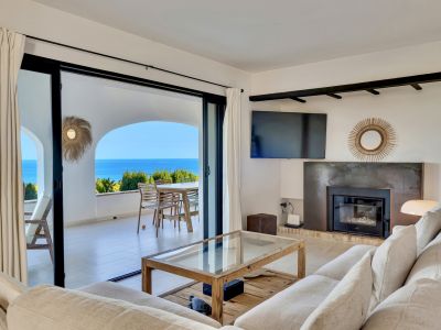 Salón luminoso con sofá claro, mesa de centro de madera y chimenea moderna. Amplios ventanales se abren a una terraza con vistas panorámicas al mar.