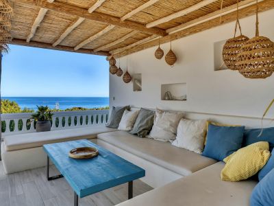 Terraza cubierta con asientos integrados, cojines variados y una mesa de madera azul. El techo de caña natural complementa las vistas panorámicas al mar Mediterráneo.