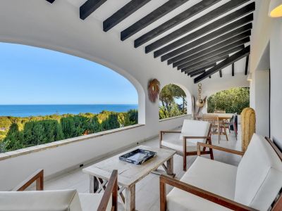Terraza cubierta con vigas de madera oscura y suelo de baldosas blancas. Ofrece vistas panorámicas al mar a través de arcos, con mobiliario de exterior en madera.