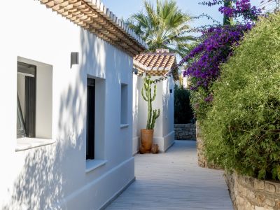 Un sendero exterior de tablas claras flanqueado por edificios blancos con tejados de teja y vegetación mediterránea, incluyendo buganvillas moradas y un cactus alto.