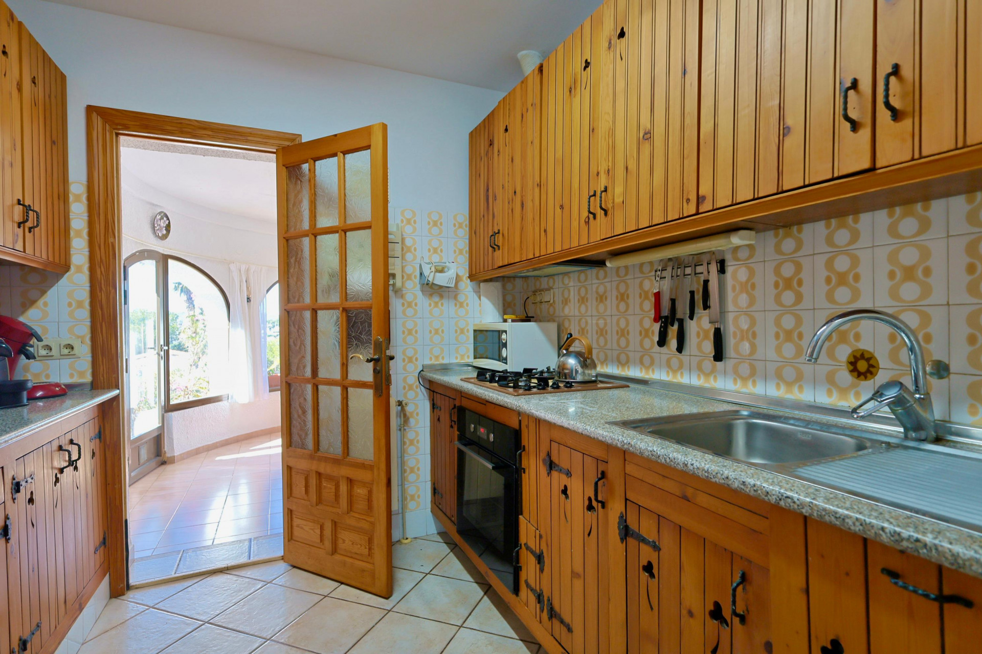 Cocina rústica con muebles de madera y acceso a un salón luminoso con ventanales.
