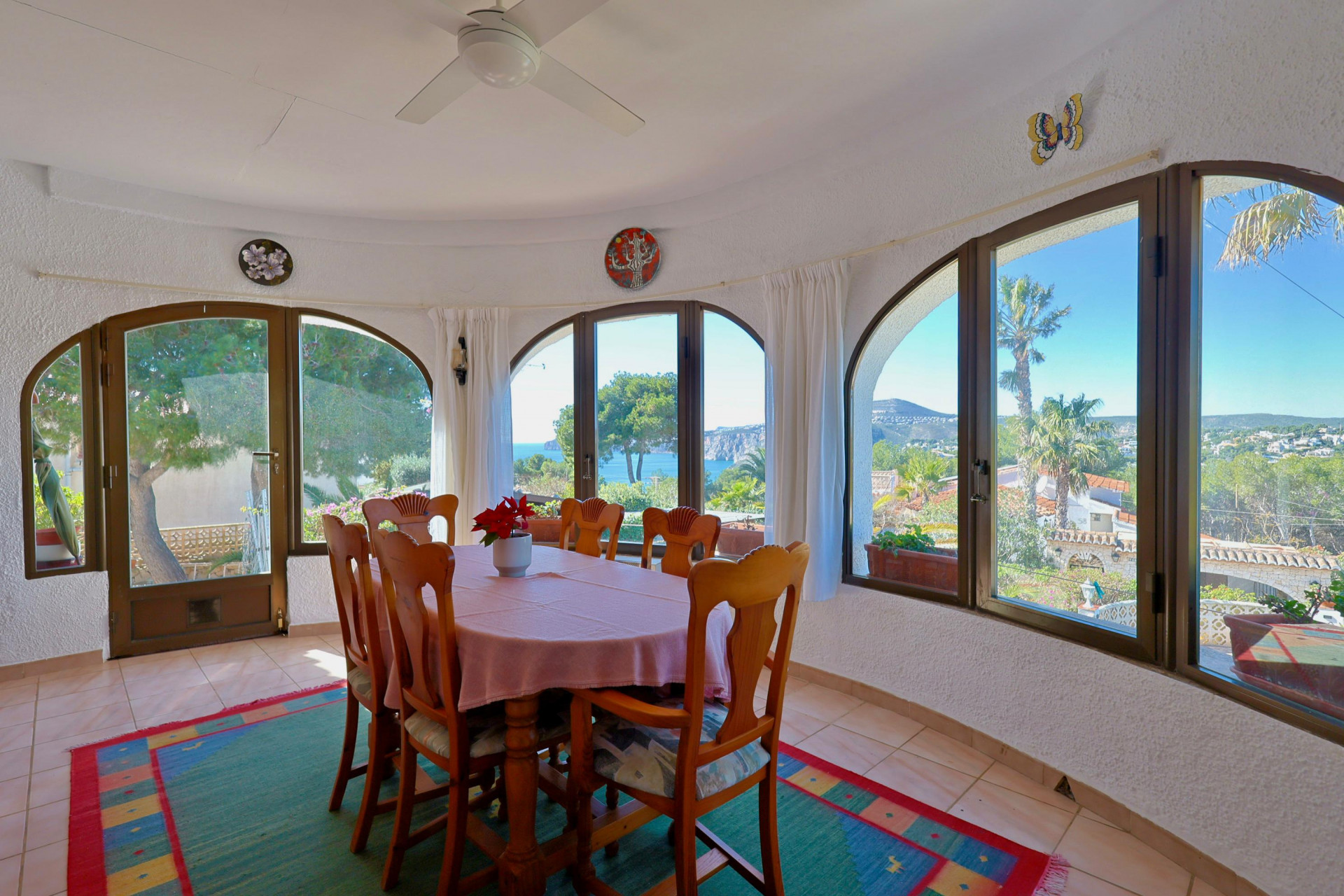Comedor luminoso con vistas al mar a través de ventanales arqueados. Mesa de madera con mantel rosa.
