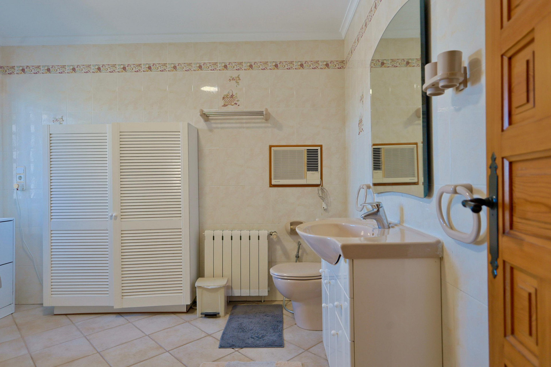 Cuarto de baño alicatado con armario de persiana blanca y lavabo con encimera beige.