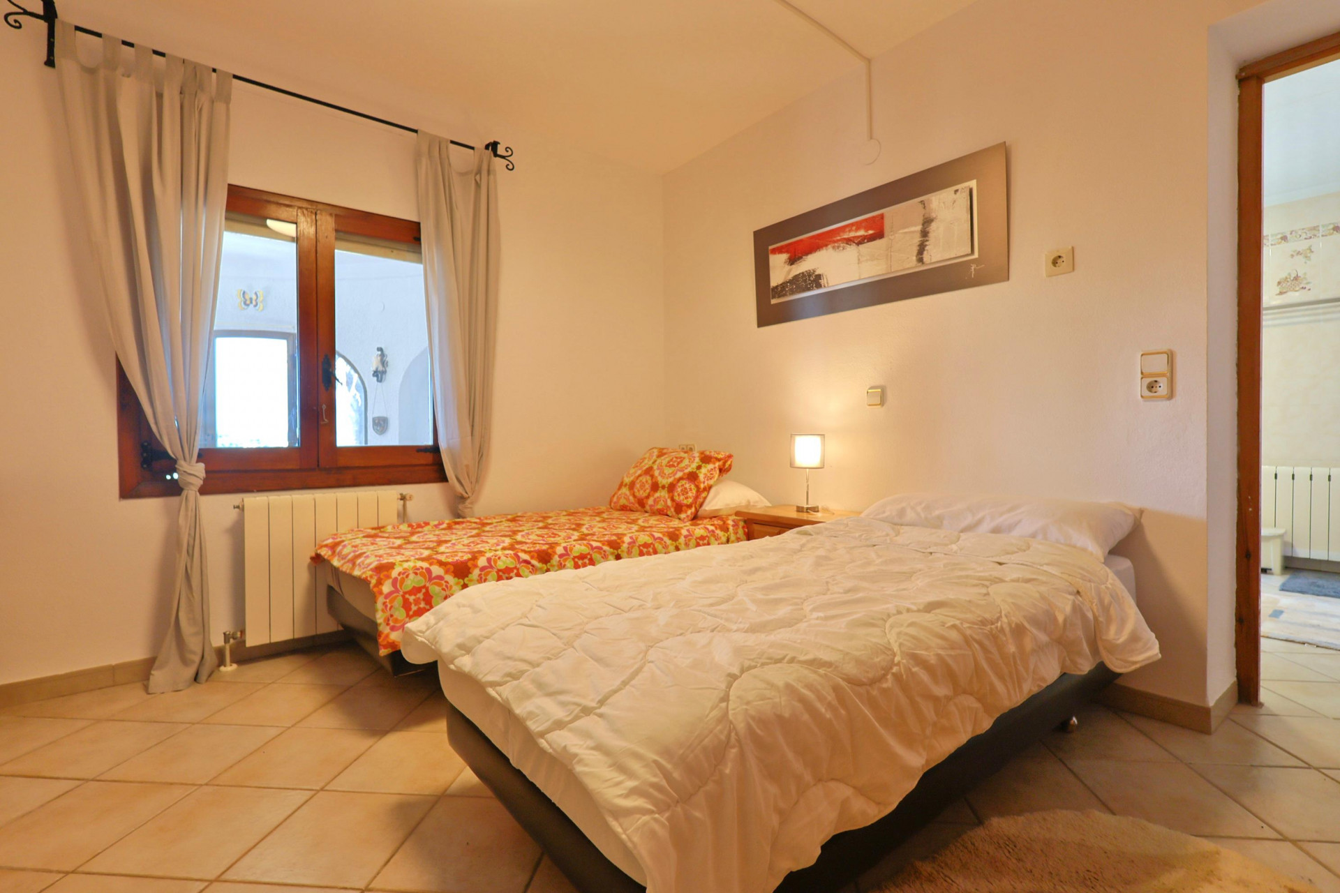 Dormitorio luminoso con dos camas individuales, ventana de madera y suelo de baldosas.