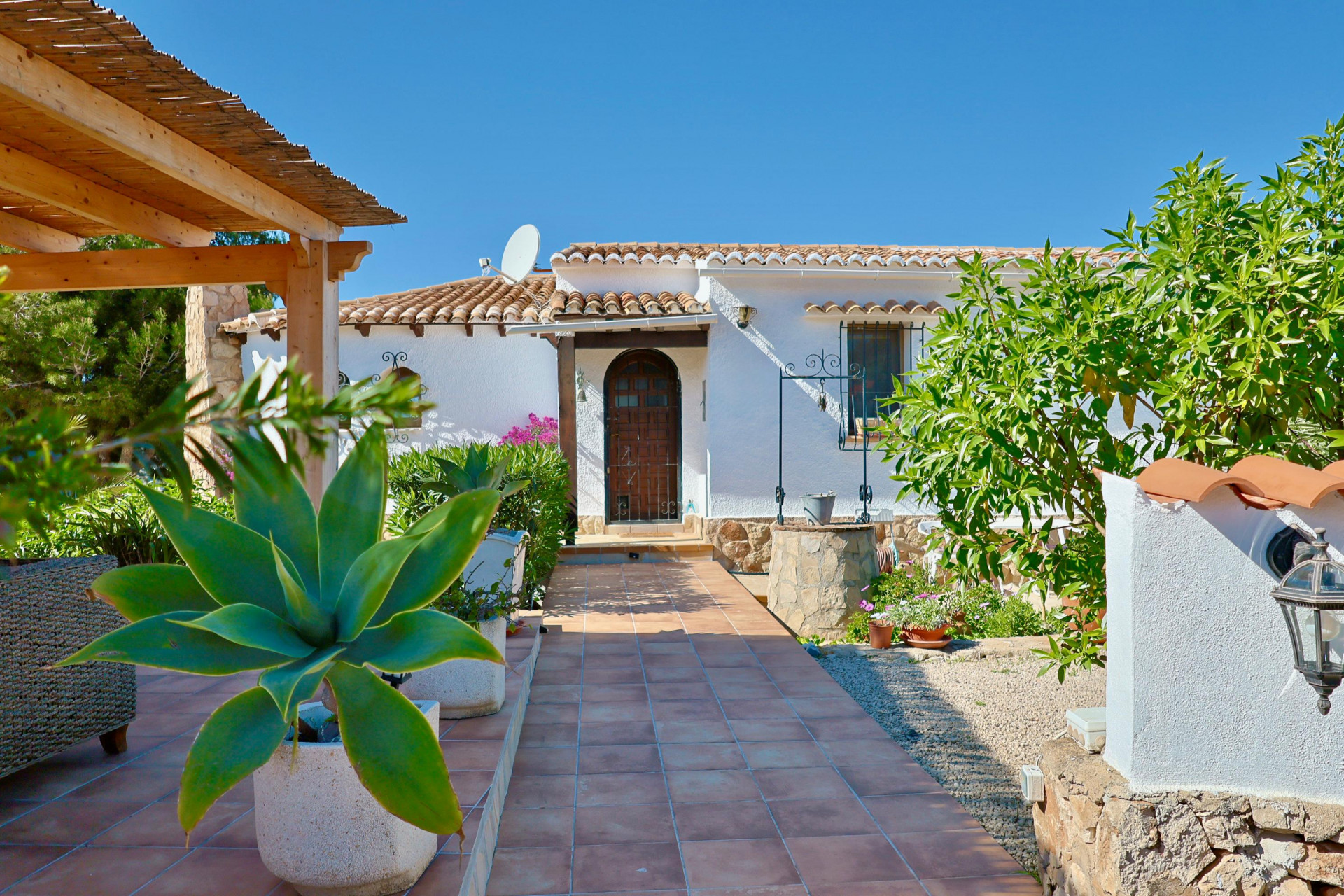 Entrada a la villa con camino de baldosas, vegetación mediterránea y pozo de piedra.