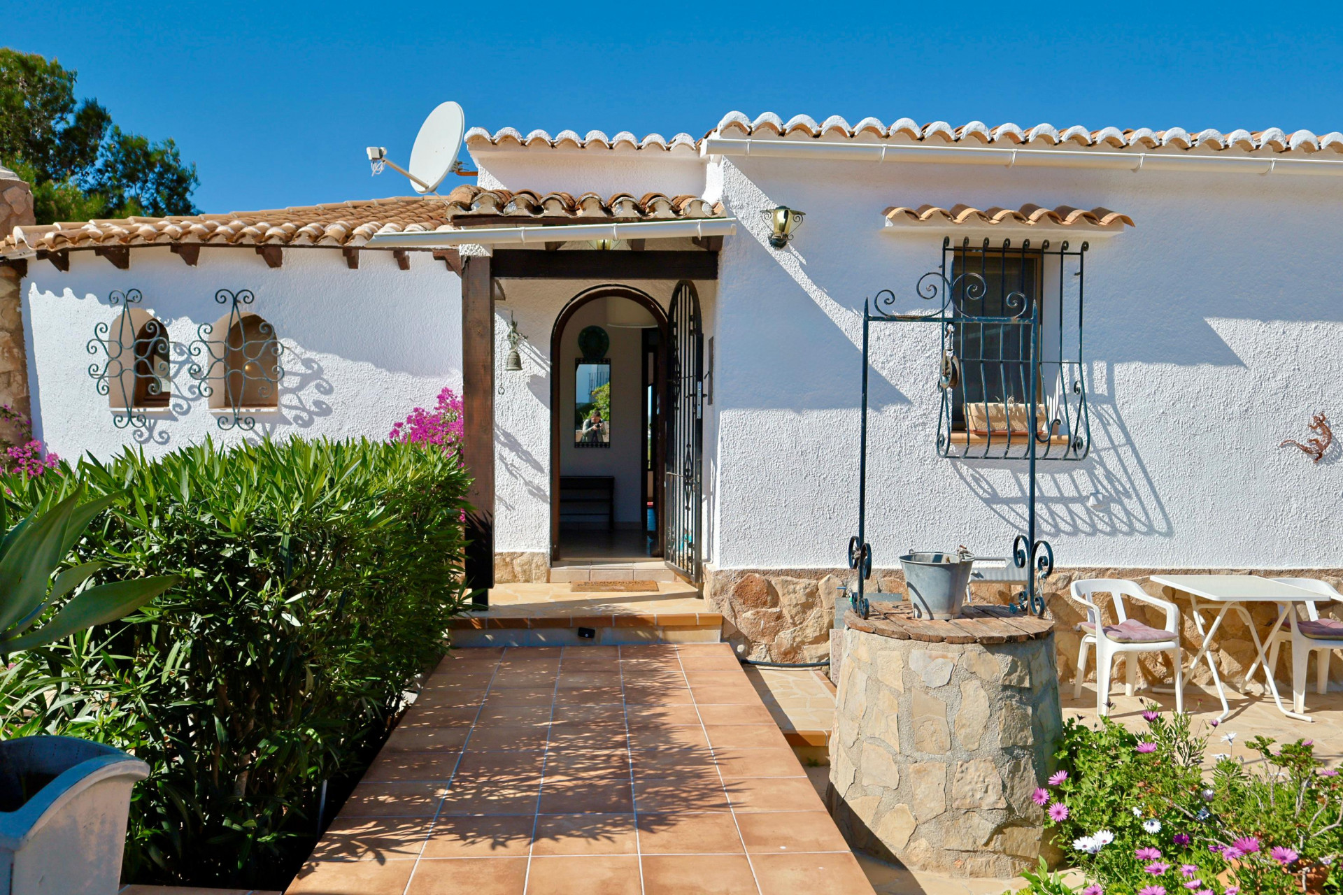 Entrada a la villa con pozo de piedra, camino de baldosas y fachada blanca texturizada.
