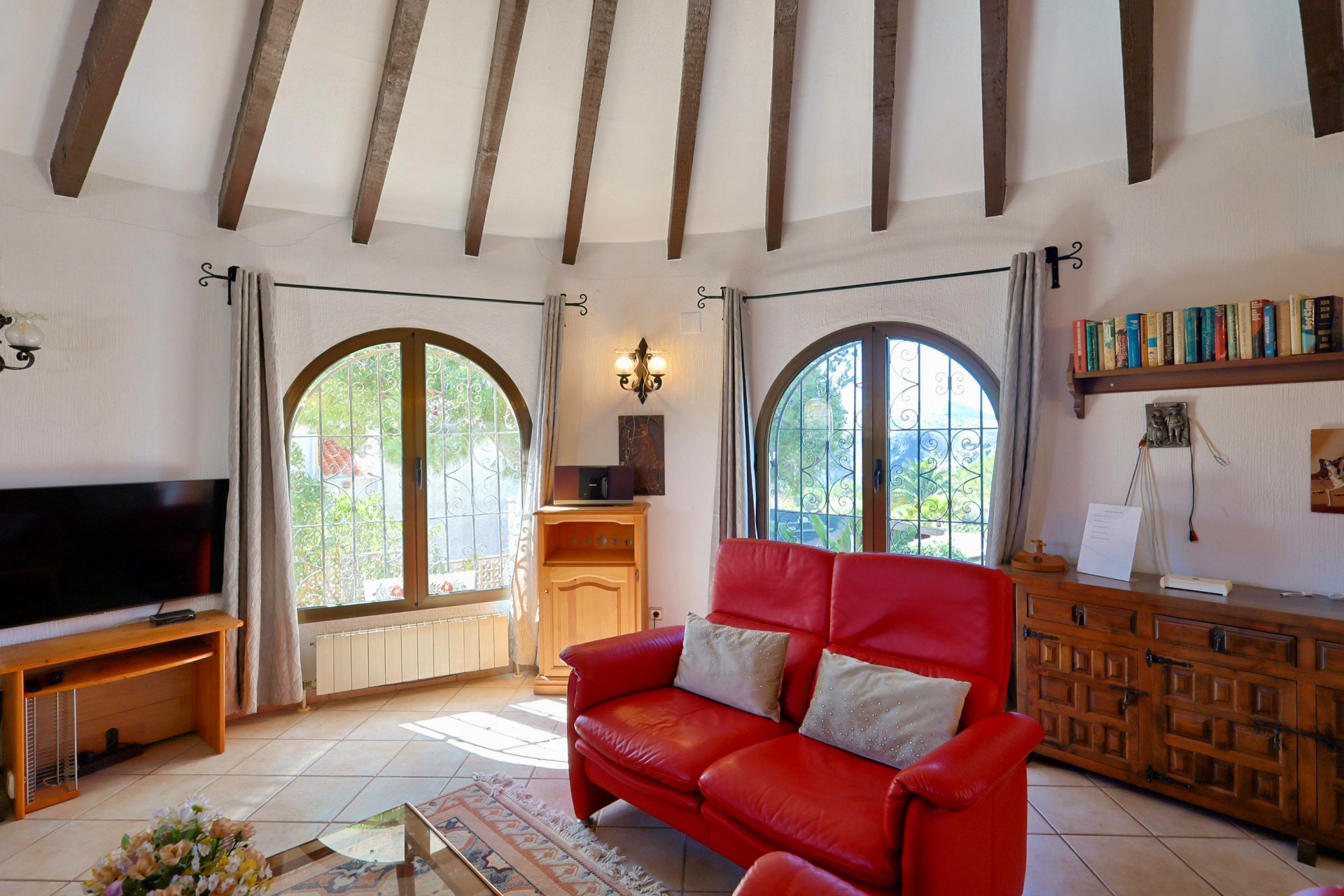 Salón luminoso con vigas de madera, sofá de cuero rojo y ventanas arqueadas con vistas al jardín.