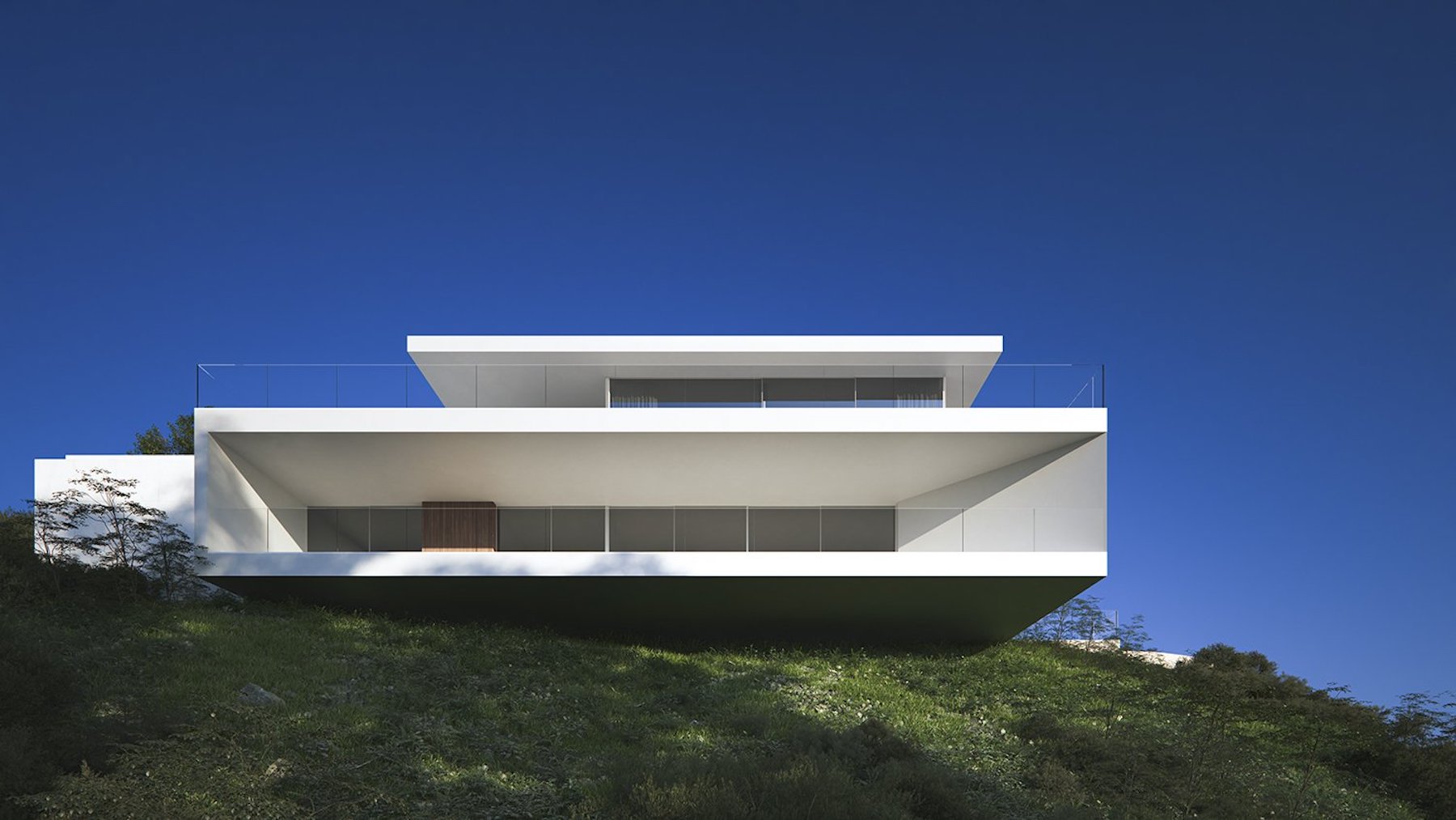 Fachada exterior de diseño minimalista con voladizos geométricos blancos y barandillas de vidrio, integrada en una ladera natural bajo un cielo despejado.