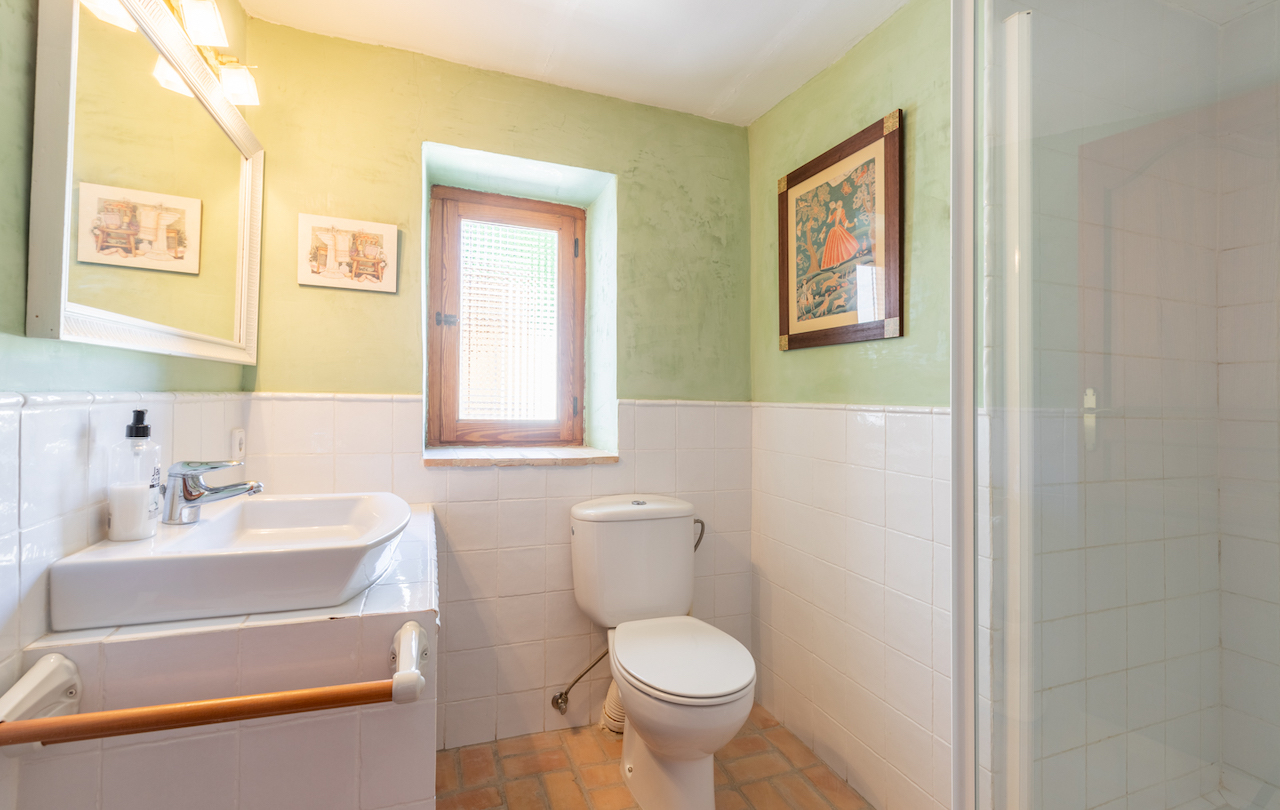 Baño de estilo rústico con paredes en verde suave, azulejos blancos, suelo de barro cocido y una ventana de madera que aporta luz natural.