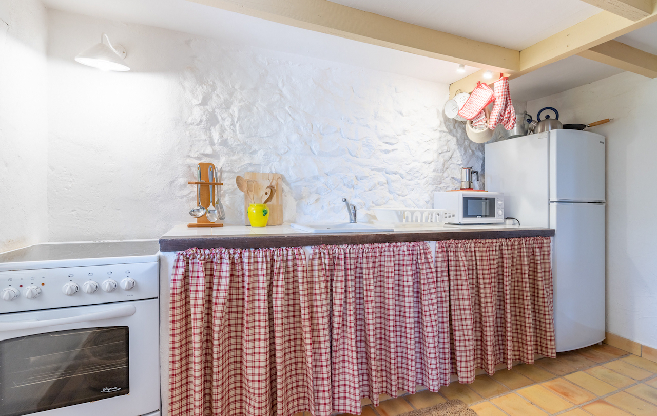 Cocina de estilo rústico con paredes de piedra encalada, vigas de madera y suelos de barro. Incluye electrodomésticos blancos y encimera con cortinas de cuadros tradicionales.