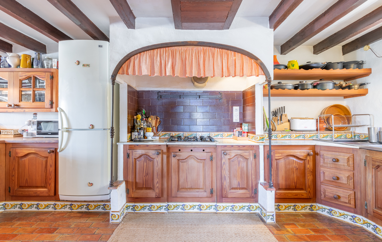 Cocina de estilo tradicional valenciano con vigas de madera vista, suelos de barro cocido y zona de cocción bajo un arco decorativo con azulejos ornamentados.