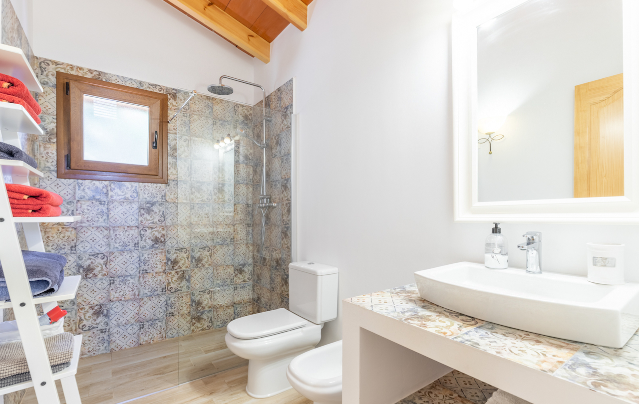 Cuarto de baño con techo de vigas de madera, ducha italiana con azulejos hidráulicos y encimera a juego con lavabo sobrepuesto y sanitarios blancos.