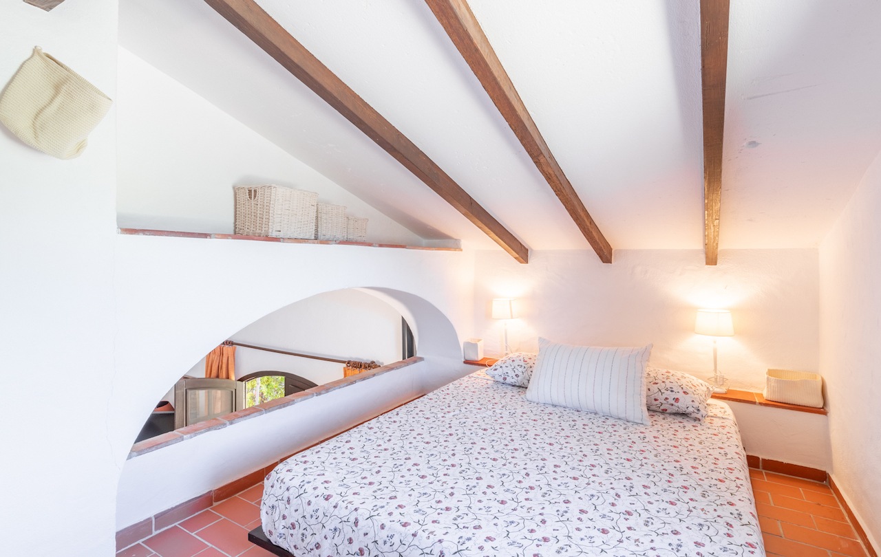Dormitorio abuhardillado con vigas de madera vistas, suelos de barro cocido y un arco arquitectónico que comunica visualmente con la estancia inferior.