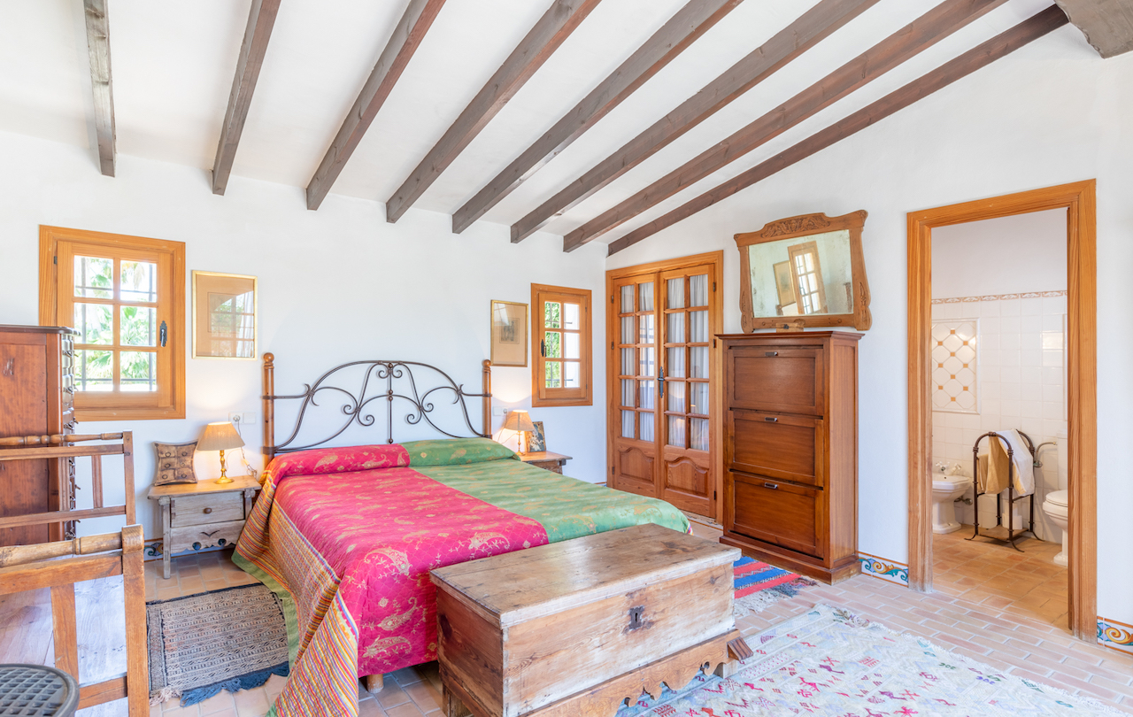 Dormitorio rústico con vigas de madera a la vista, suelos de barro cocido y mobiliario tradicional. Incluye acceso directo a un baño en suite y techos inclinados.