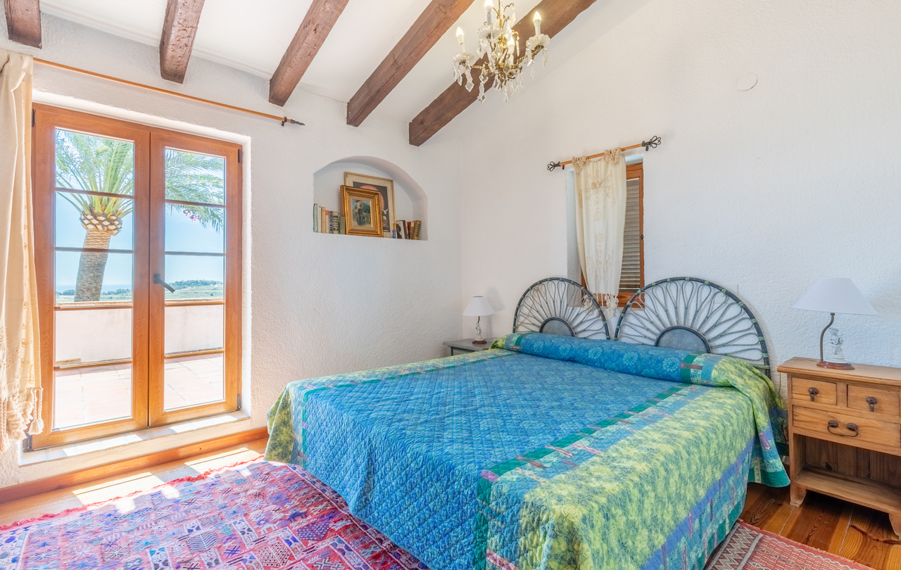 Dormitorio rústico con vigas de madera vistas, nicho decorativo en pared y acceso directo a terraza con vistas a palmeras. Incluye suelos de madera y cabecero de forja.