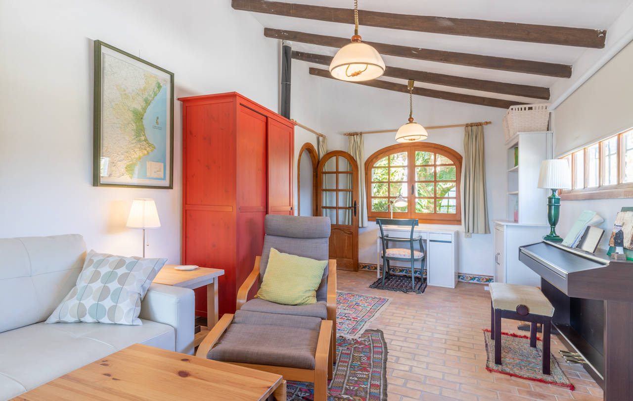 Sala de estar con vigas de madera vista, suelos de barro cocido y un gran armario rojo. Incluye zona de estudio bajo ventana de arco y un piano digital.