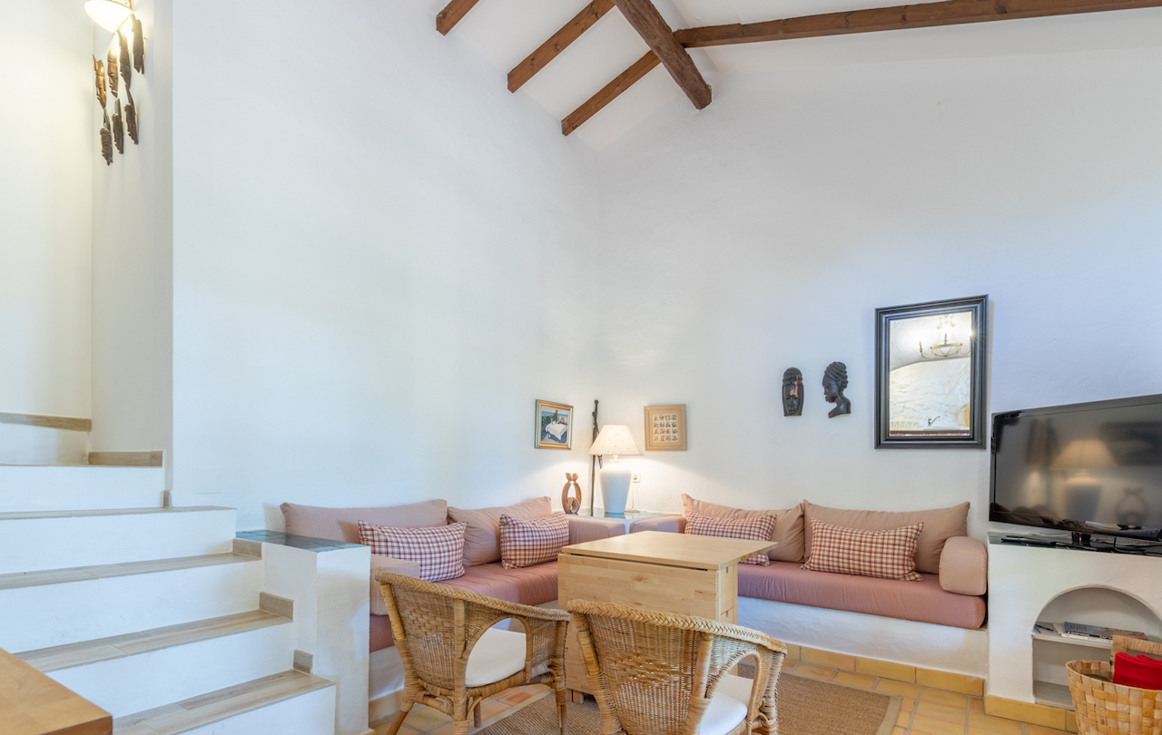 Salón de estilo rústico con techos altos y vigas de madera vistas. Incluye sofás de obra, mobiliario de mimbre, suelos de barro cocido y acceso por escalera lateral.