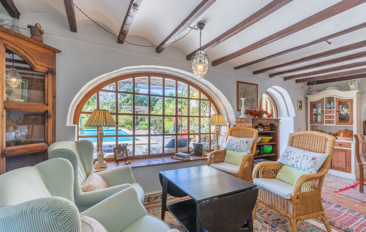 Salón de estilo tradicional con vigas de madera vistas, suelos de barro y un gran ventanal de arco con vistas directas a la piscina y al jardín mediterráneo.