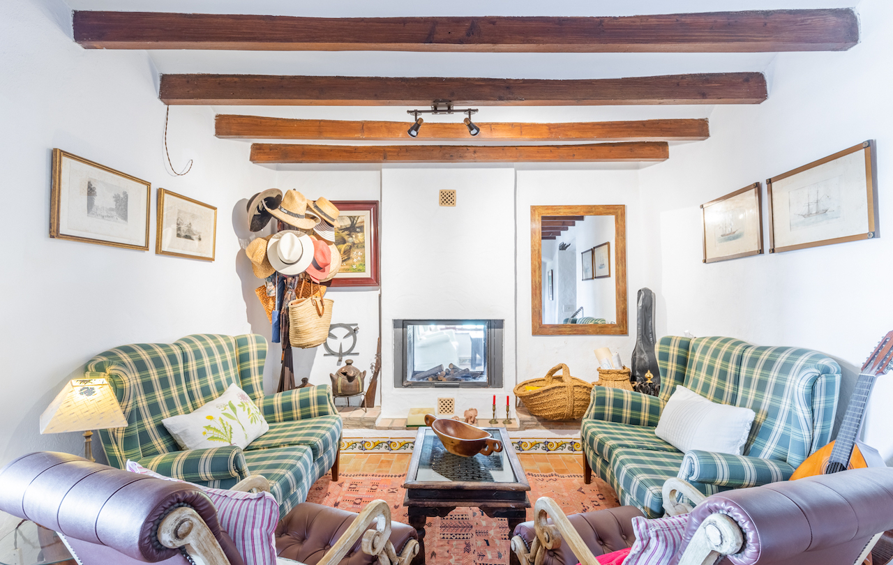 Salón rústico con vigas de madera vista, chimenea encastrada y suelos de barro cocido con cenefas cerámicas. Dispone de sillones de cuadros y mobiliario tradicional valenciano.