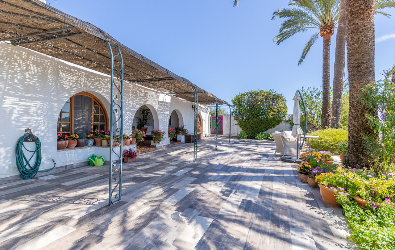 Terraza exterior con pavimento cerámico de efecto madera, pérgola de cañizo sobre pilares de forja y fachada blanca con arcos tradicionales decorados con macetas.