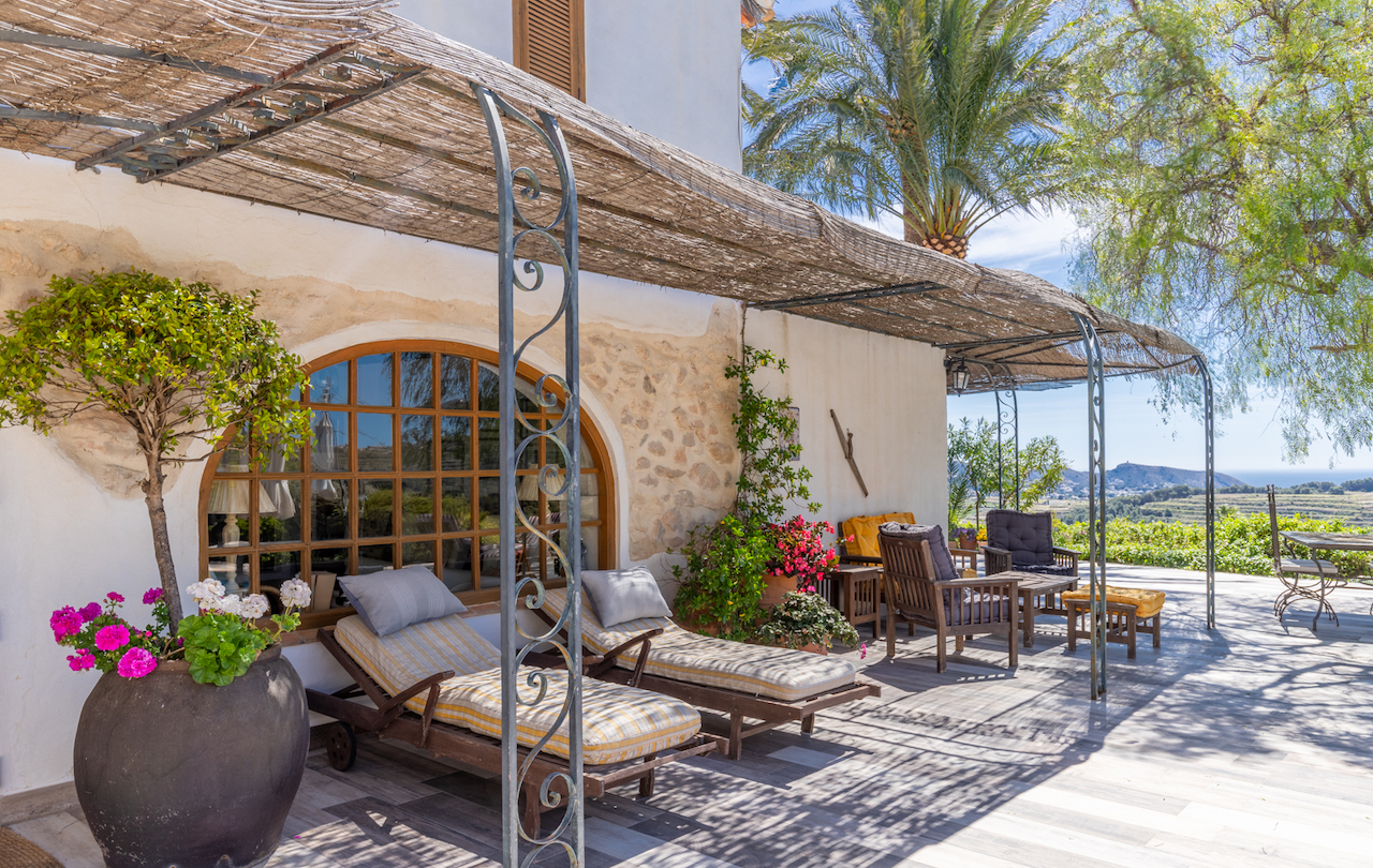 Terraza exterior con pérgola de cañizo y hierro forjado, zona de descanso con tumbonas frente a un ventanal de arco y vistas panorámicas al valle y al mar.
