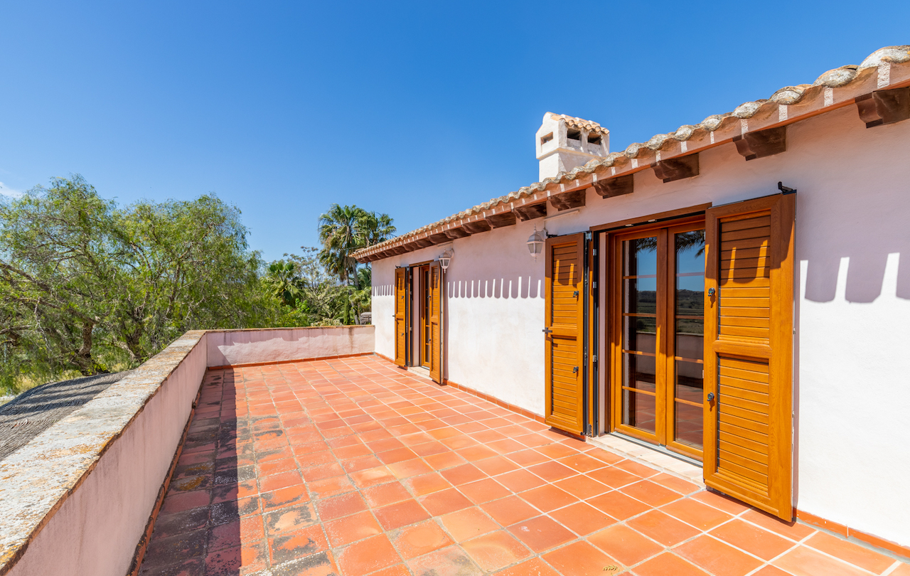 Terraza superior con suelo de baldosas de barro, vigas de madera vista bajo el tejado y puertas de madera con contraventanas mallorquinas integradas en la fachada blanca.