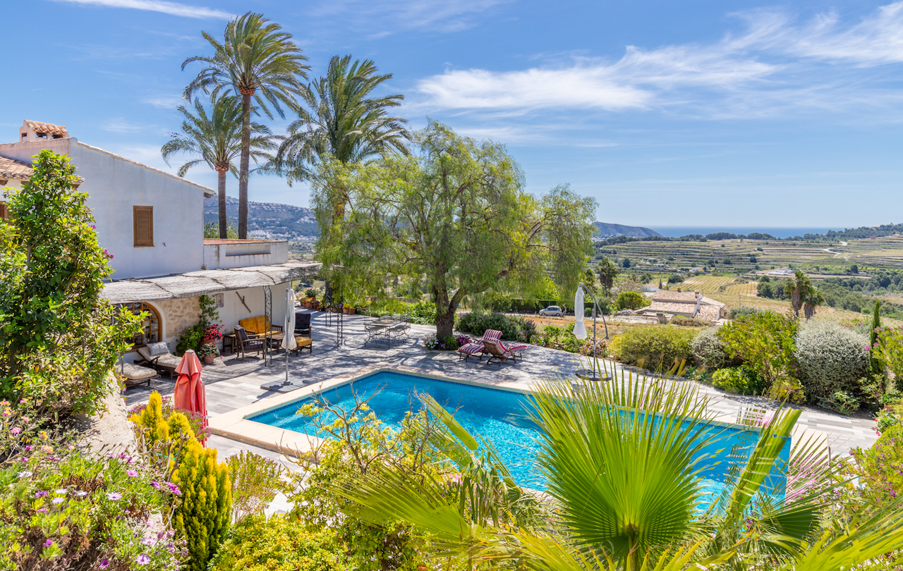 Zona de piscina con amplia terraza de piedra y jardines mediterráneos, ofreciendo vistas panorámicas hacia los campos de viñedos y el mar de Moraira.
